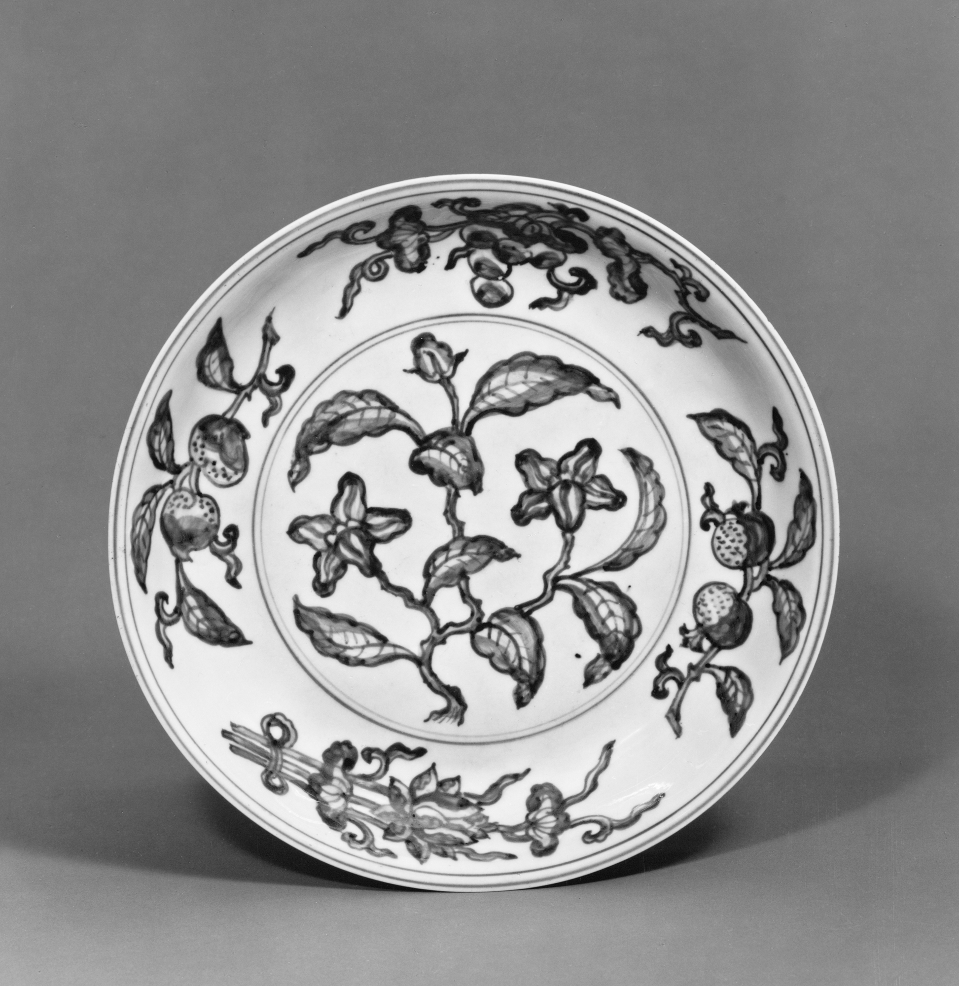 Plat à fond clair, décoré au centre de trois branches fruitières — pêche, grenade, raisin — disposées en composition rayonnante.